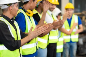 Group of workers in hard hats and reflective vests clapping together, in celebration.